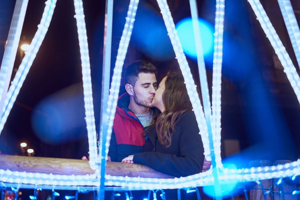 Una pareja se besa en el centro de la imagen mientras les rodean las luces blancas y azules de una feria. 