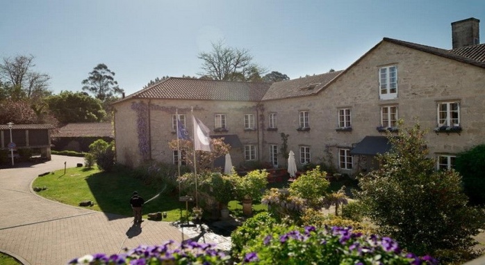 Foto de la fachada de A Quinta da Auga, rodeada de plantas, flores y un hórreo. Siente Galicia.
